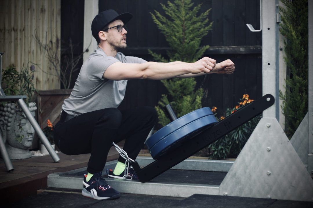 Photo 09-03-2022, 17 42 53 man performing a belt squat with the forte fitness belt squat attachment with 2 blue 20kg plates, on an outdoor rig surrounded by trees