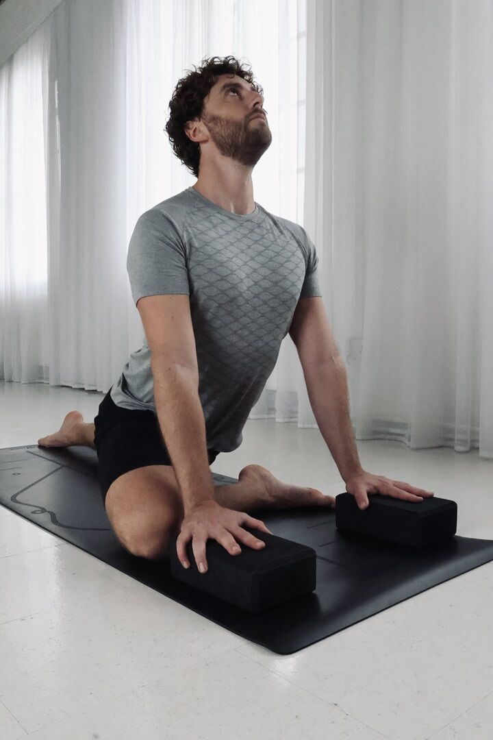 yoga block man performing a pigeon pose on a black yoga mat, using some black foam yoga blocks props