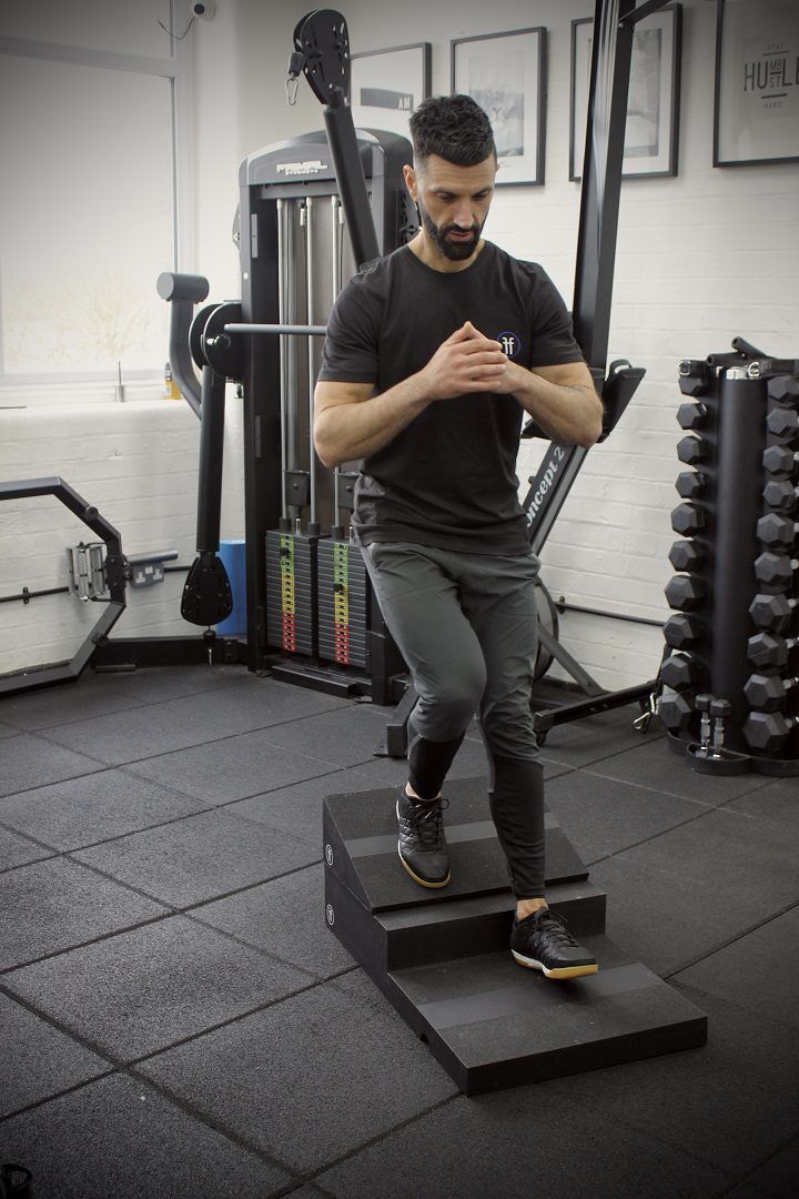 poliquin step A man using the Forte Fitness step and incline boards. The incline board is placed on the top step, and the man is at the beginning of his squat, with his right leg placed on the incline board.