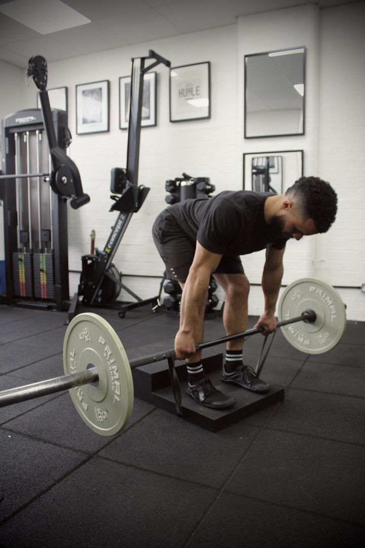 banded deadlift A man using the Forte Fitness step. He is stood on the lower step, and is bending over in a forward position while holding a bar with weights attached. His knees are bent to complete the deadlift maneuver.