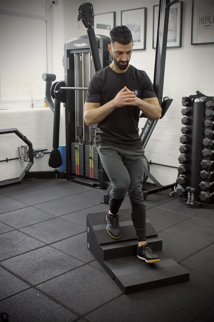 poliquin step A man using the Forte Fitness step and incline boards. The incline board is placed on the top step, and the man is at the beginning of his squat, with his right leg placed on the incline board.